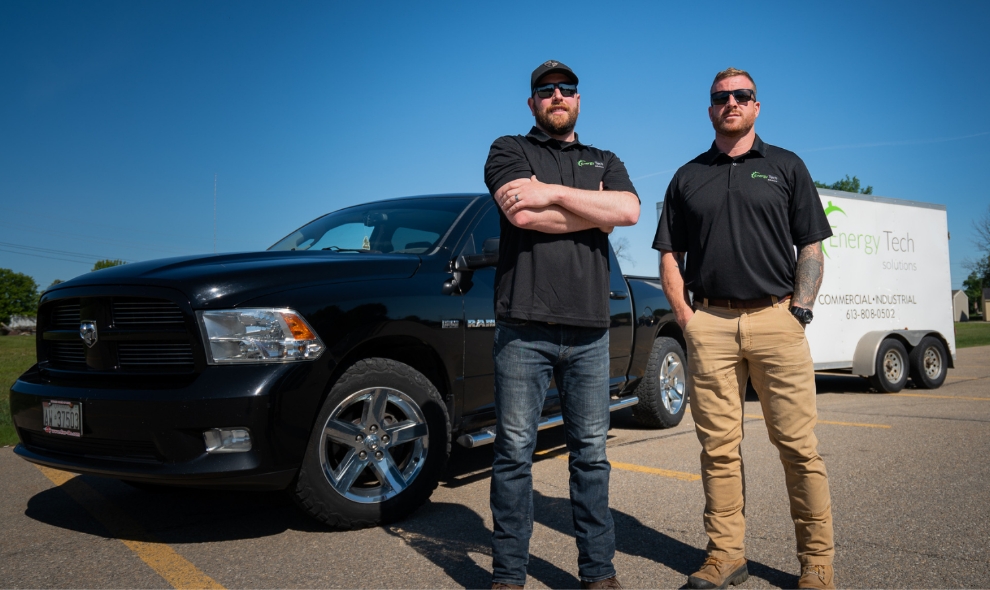 Two males standing in front of their black truck and Energy Tech trailer