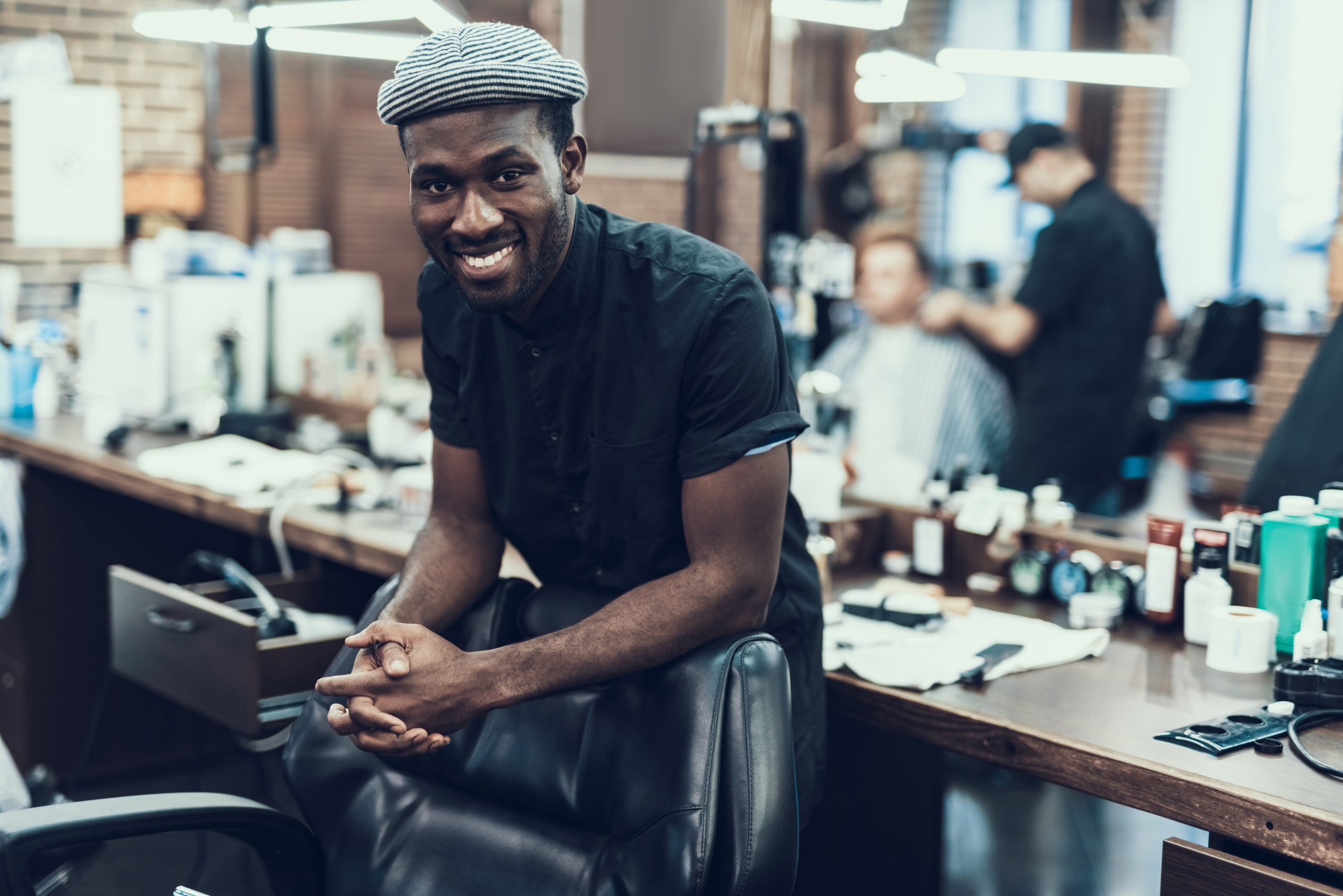 A smiling barber stands in a barbershop, with client reflected in mirror behind him