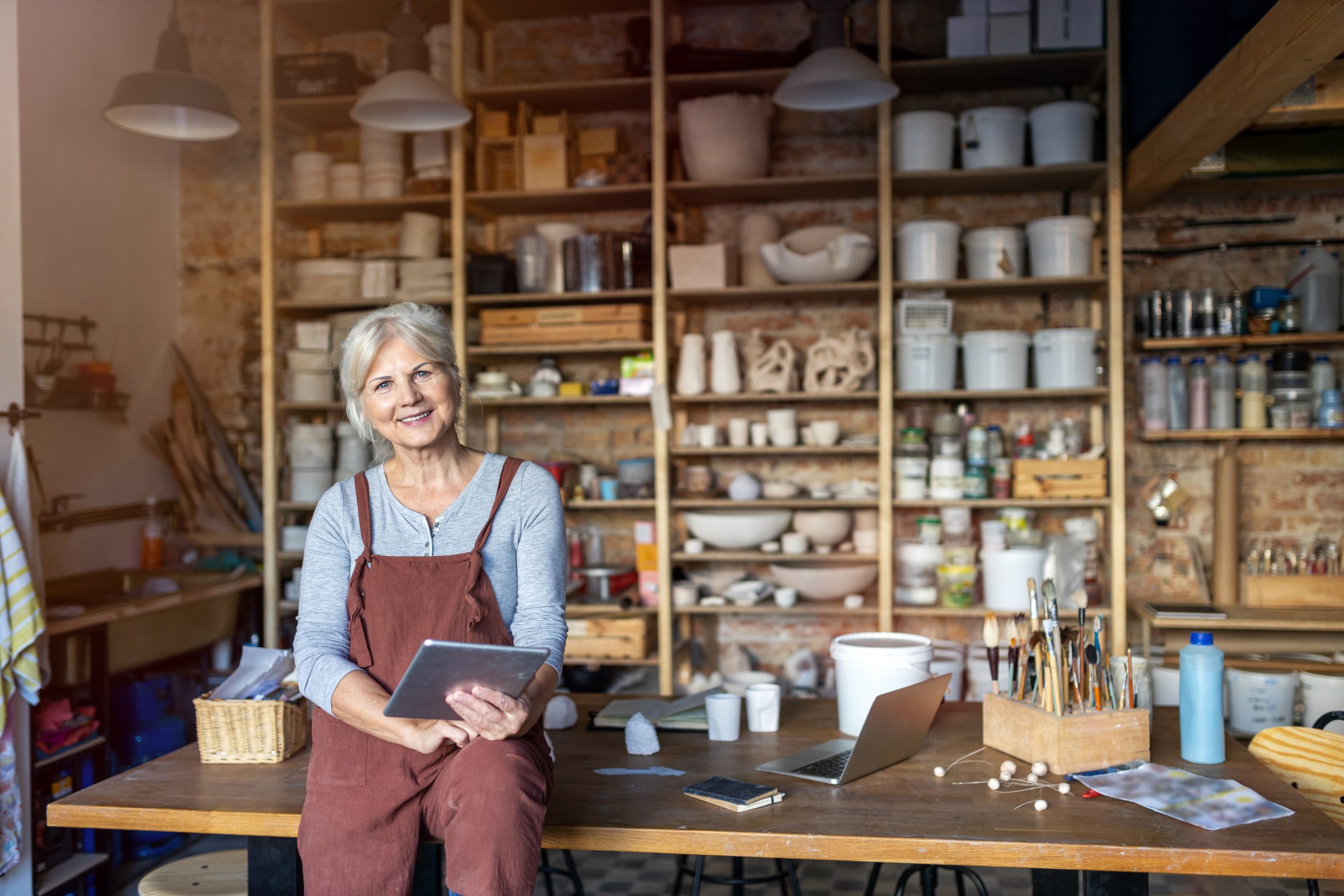 Older woman looks at the camera, leaning against her craft table with ceiling high shelves of pottery behind her