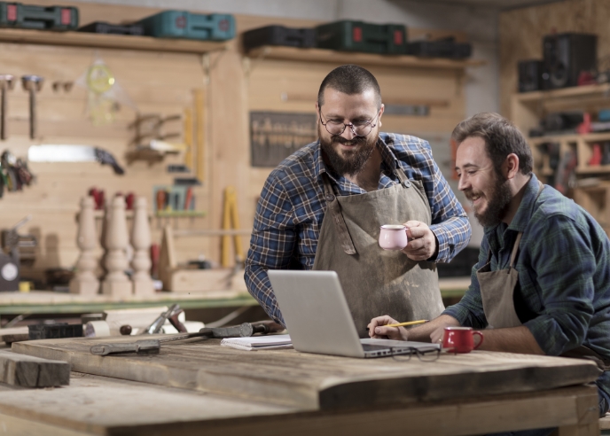 Two woodsman looking at a laptop while enjoying coffee in their wood shop